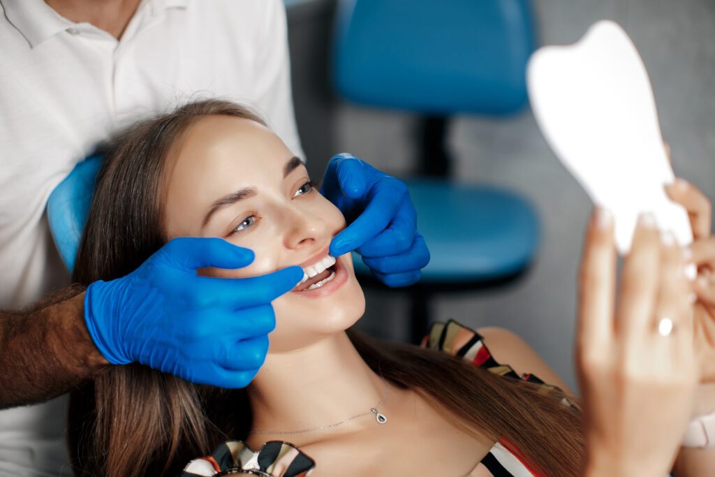 young woman observing her bonded teeth in the mirror, with a doctor in blue glove in the background
